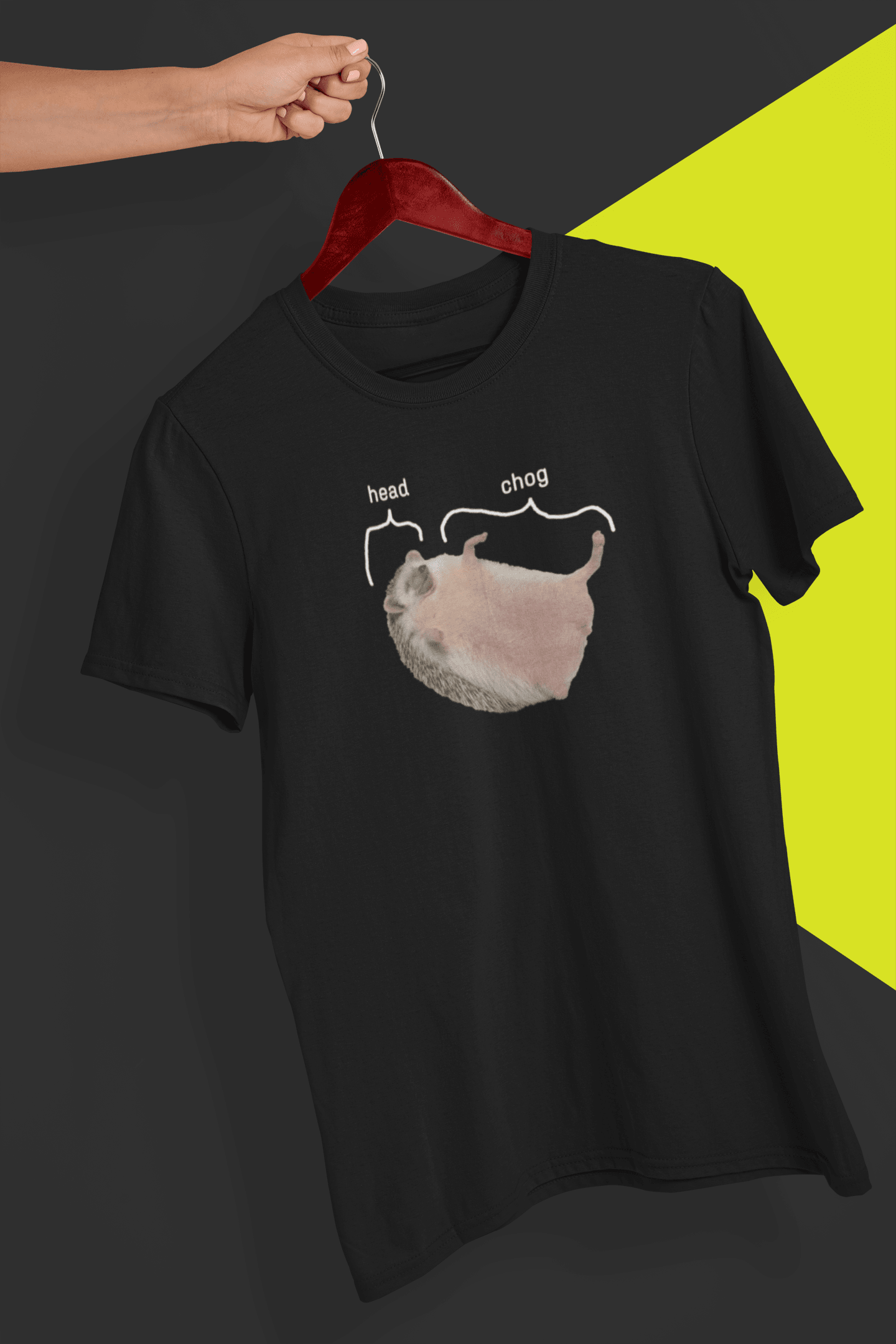 A black T-shirt featuring a cute hedgehog, which says &quot;head&quot; pointing at his head, and &quot;chog&quot; pointing at the rest of his body, is hung on a red hanger, held by a hand against a split black and yellow background.