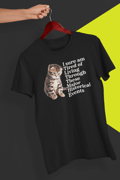 A black T-shirt with a cute kitten written &quot;I sure am Tired of Living Through These Major Historical Events;. is hung on a red hanger, held by a hand against a split black and yellow background.