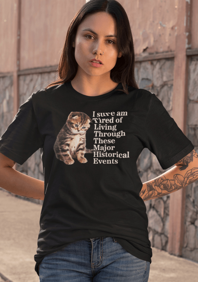 A woman wears a black T-shirt featuring a cute sad cat written &quot;I sure am Tired of Living Through These Major Historical Events&quot;.