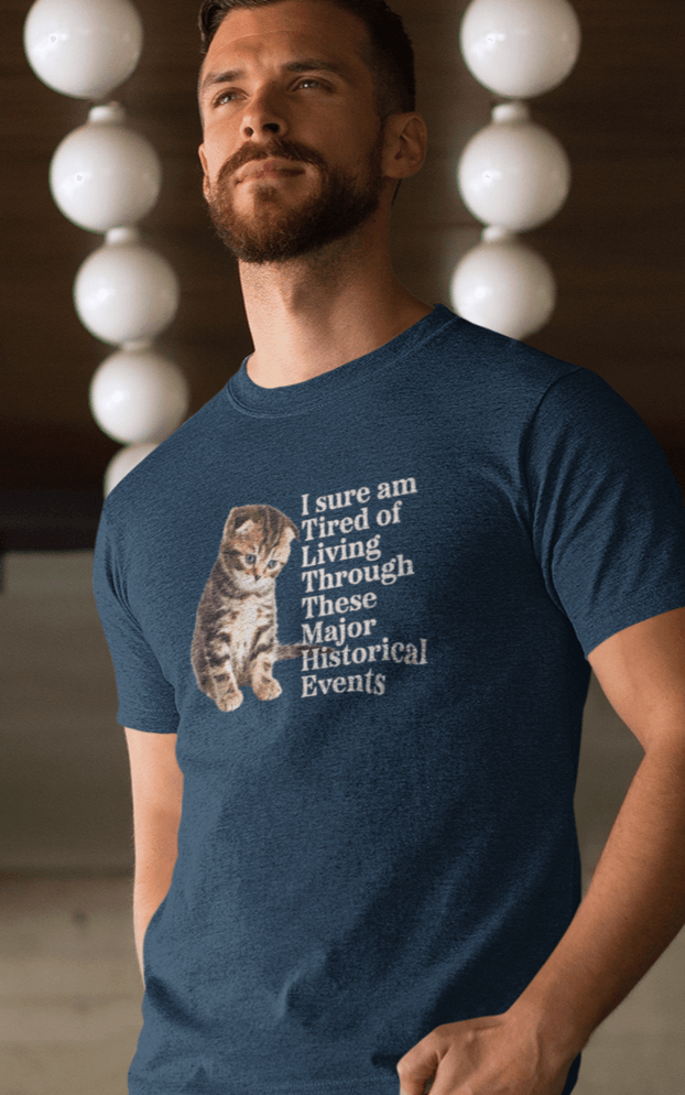 A man wears a navy blue T-shirt featuring a cute sad cat written &quot;I sure am Tired of Living Through These Major Historical Events&quot;.