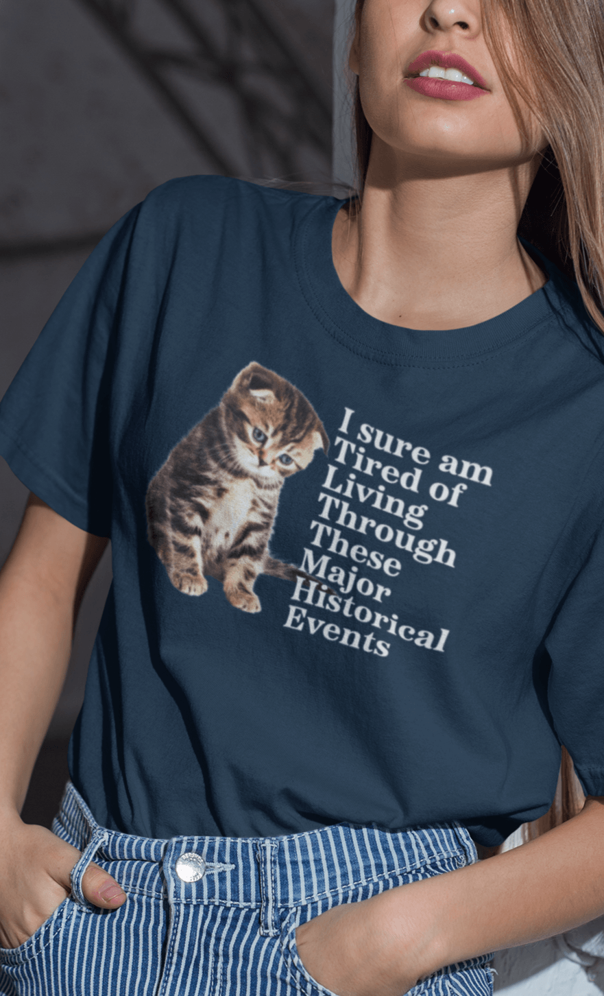 A woman wears a navy blue T-shirt featuring a cute sad cat written &quot;I sure am Tired of Living Through These Major Historical Events&quot;.