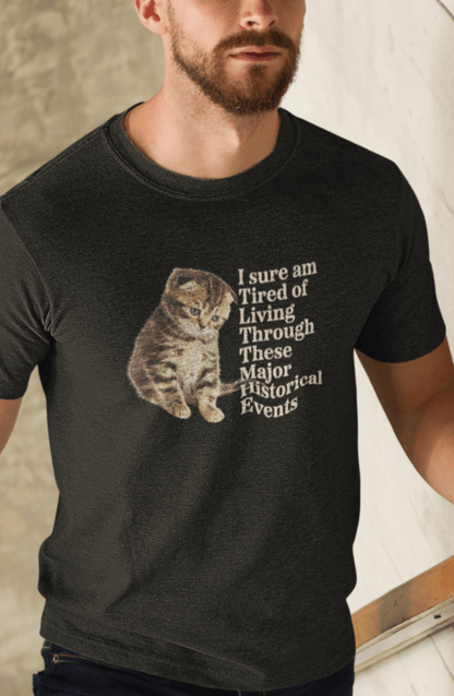 A man wears a black T-shirt featuring a cute sad cat written &quot;I sure am Tired of Living Through These Major Historical Events&quot;.