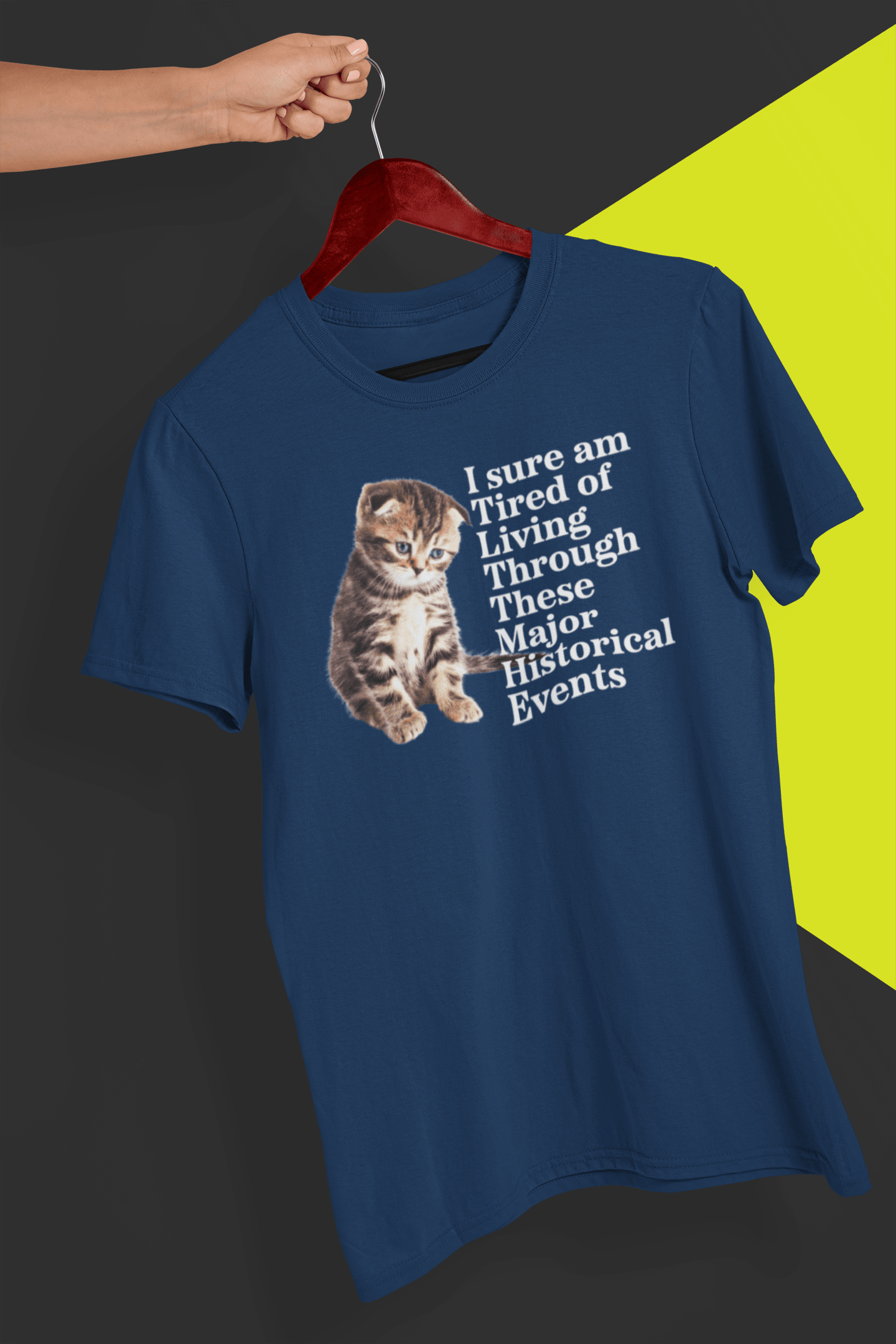 A navy blue T-shirt with a cute kitten written &quot;I sure am Tired of Living Through These Major Historical Events;. is hung on a red hanger, held by a hand against a split black and yellow background.