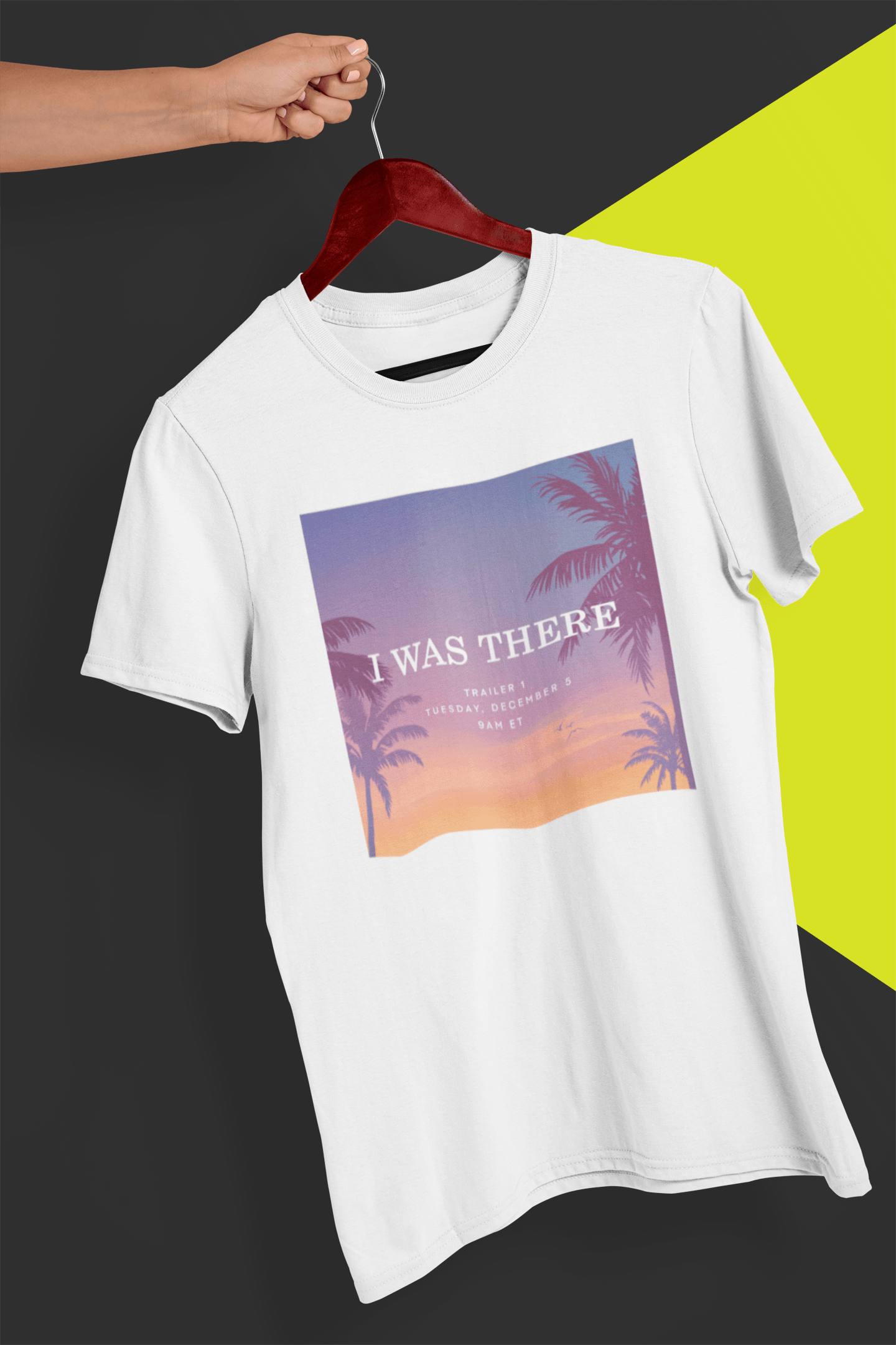 A white T-shirt with an aesthetic miami imagery written &quot;I WAS THERE, Trailer 1, Tuesday, December 5, 9AM ET&quot;, held by a hand against a split black and yellow background.