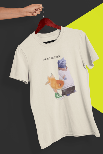 An off-white T-shirt with a cute cat and fish portraying Gumball and Darwin with &quot;me af as fuck&quot; written on top, is hung on a red hanger, held by a hand against a split black and yellow background.