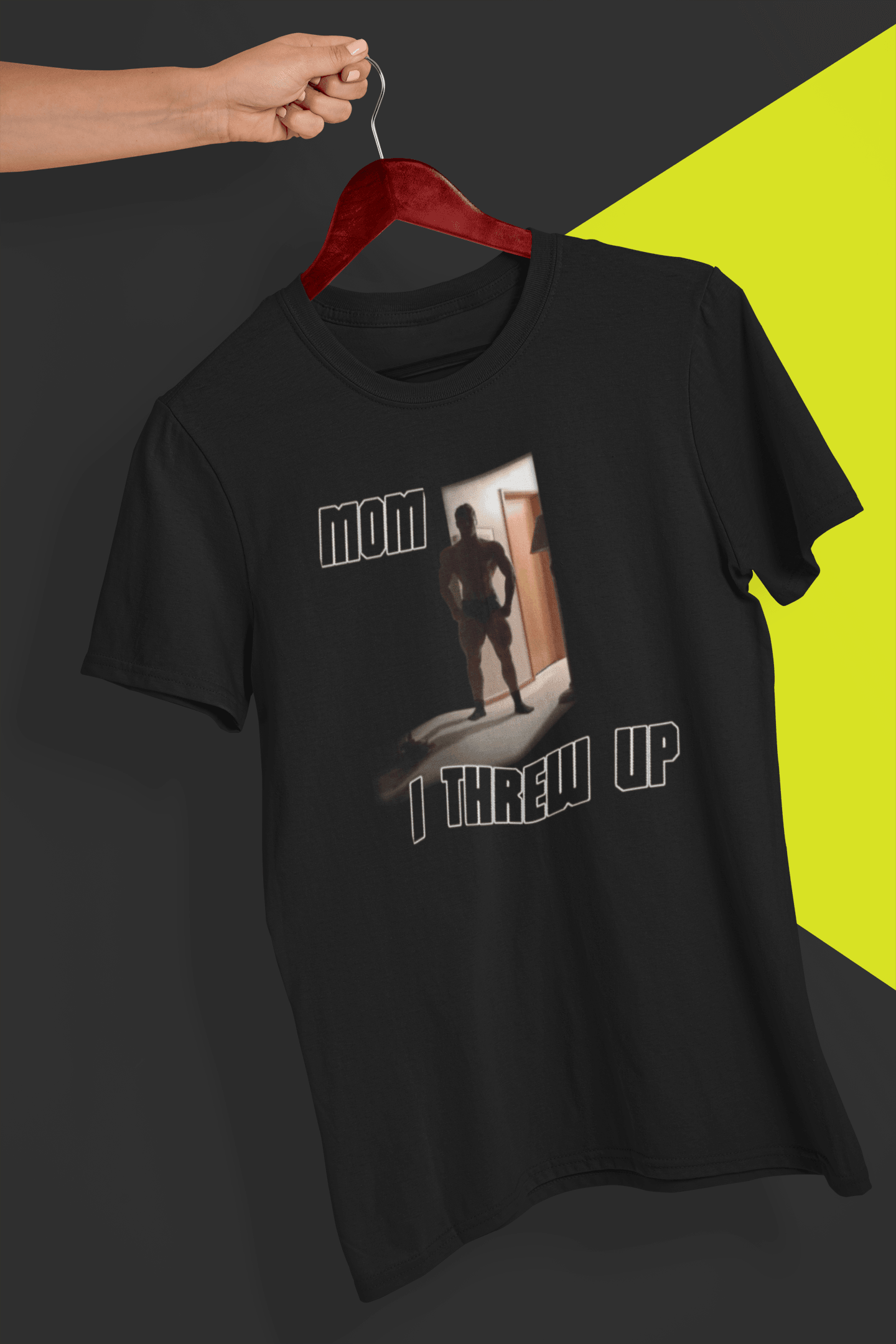 A black T-shirt with a  strong man written &quot;Mom I threw up&quot;, is hung on a red hanger, held by a hand against a split black and yellow background.