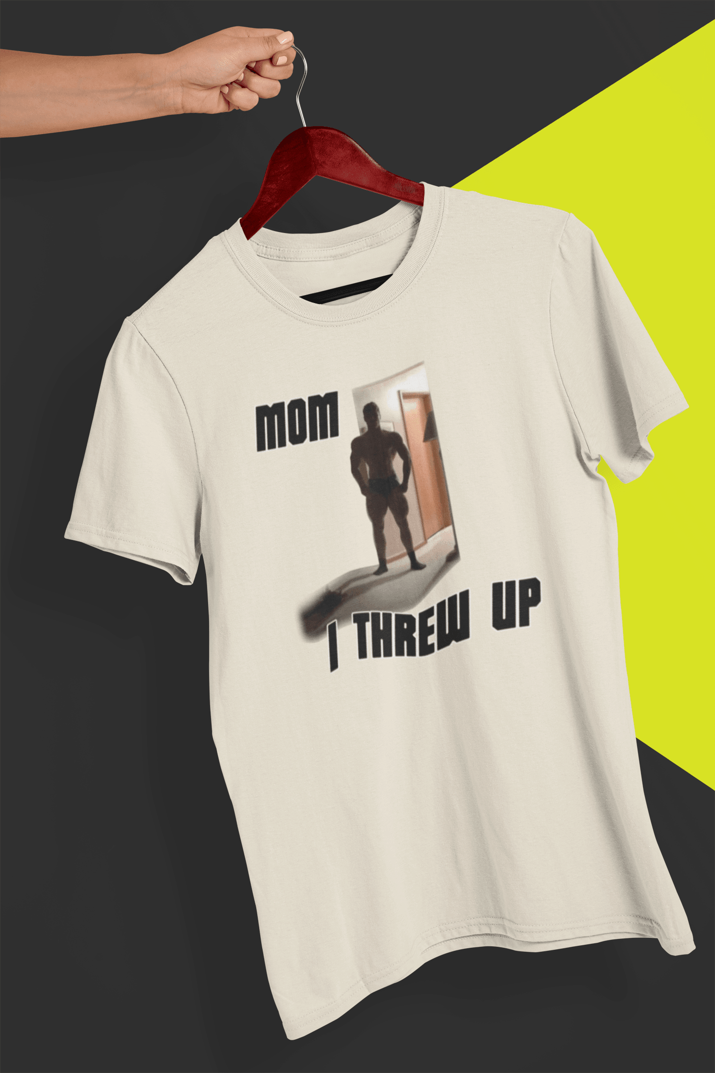 An off-white T-shirt with a  strong man written &quot;Mom I threw up&quot;, is hung on a red hanger, held by a hand against a split black and yellow background.