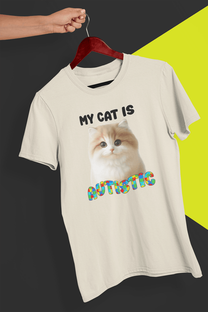 An off-white T-shirt with a cute cat written &quot;My Cat is Autistic&quot; on top, is hung on a red hanger, held by a hand against a split black and yellow background.