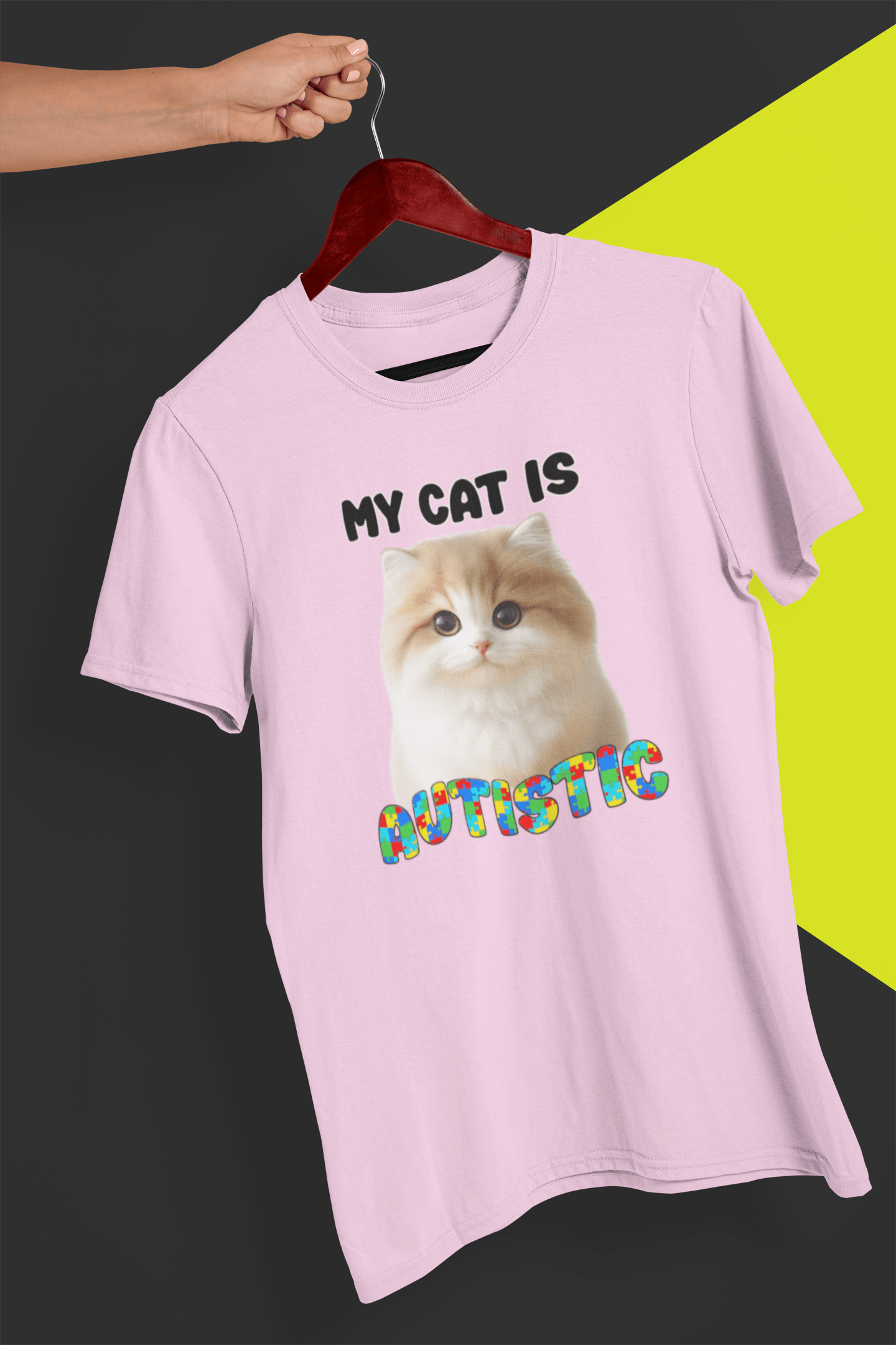 A pink T-shirt with a cute cat written &quot;My Cat is Autistic&quot; on top, is hung on a red hanger, held by a hand against a split black and yellow background.