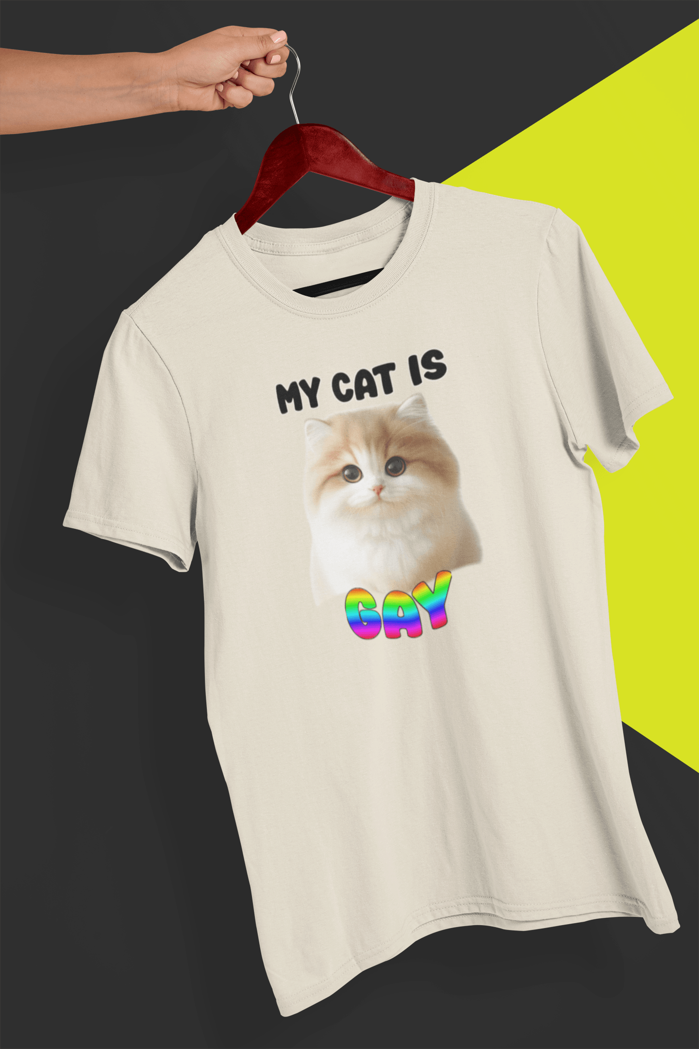 An off-white T-shirt with a cute cat written &quot;My Cat is Gay&quot; on top, is hung on a red hanger, held by a hand against a split black and yellow background.