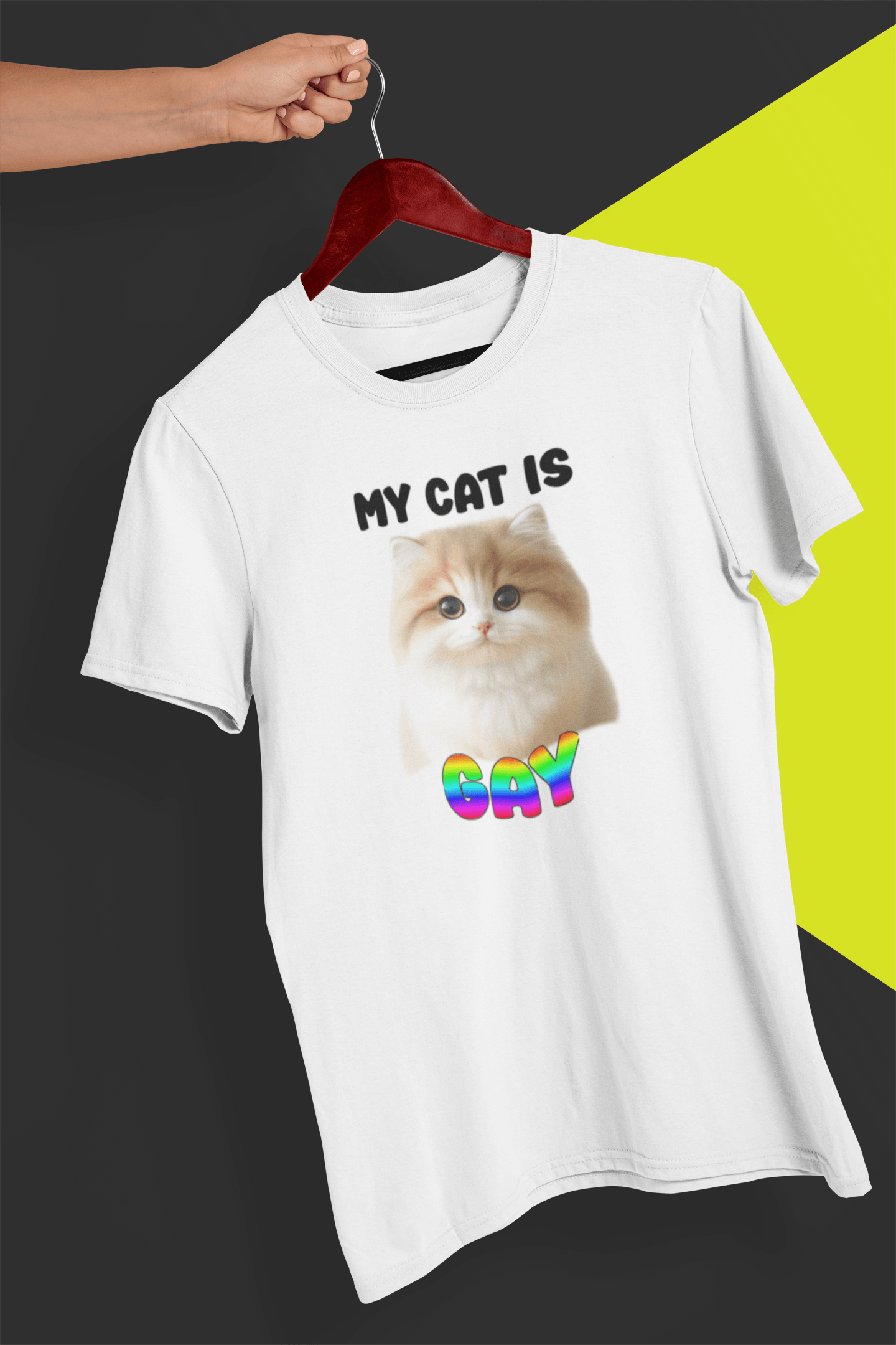 A white T-shirt with a cute cat written &quot;My Cat is Gay&quot; on top, is hung on a red hanger, held by a hand against a split black and yellow background.