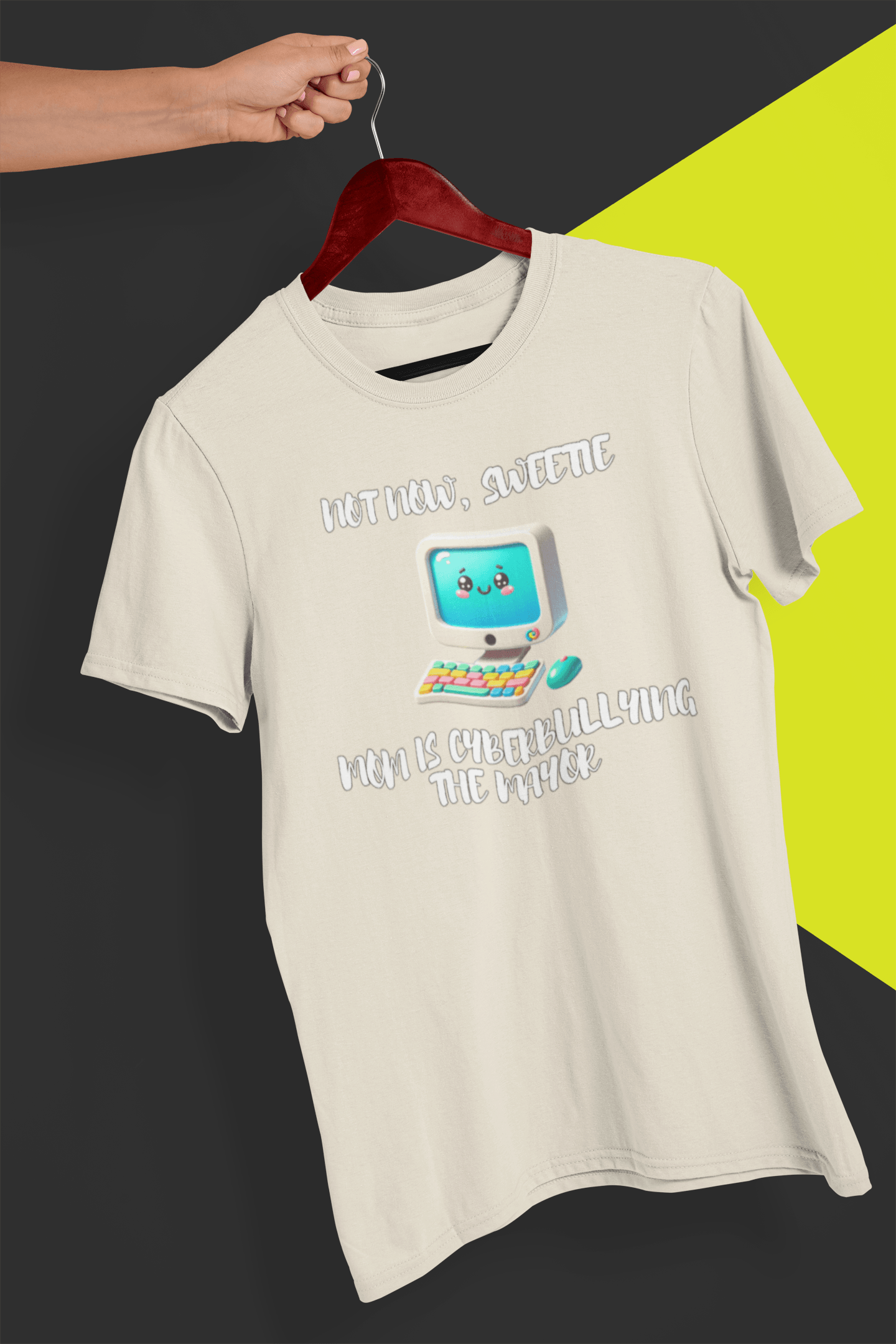 An off-white T-shirt with a cute computer written &quot;Not now, sweetie, mom is cyberbullying the mayor&quot;, held by a hand against a split black and yellow background.