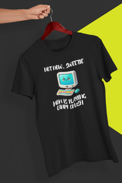 A black T-shirt with a cute computer written &quot;Not now, sweetie, mom is playing candy crush&quot;, held by a hand against a split black and yellow background.