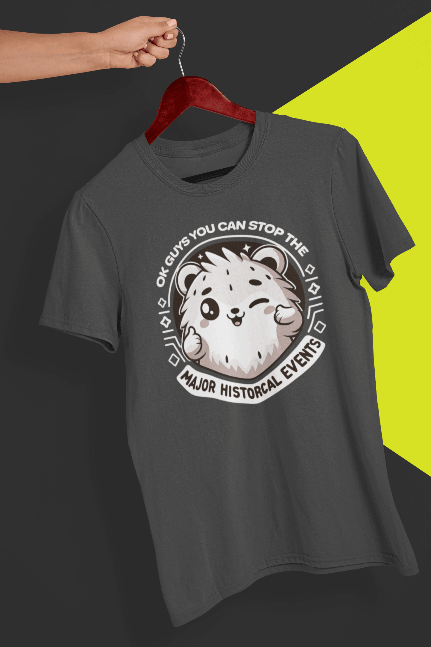 A gray T-shirt with a cute hamster doing a thumbs up written &quot;OK GUYS YOU CAN STOP THE MAJOR HISTORICAL EVENTS&quot;. is hung on a red hanger, held by a hand against a split black and yellow background.