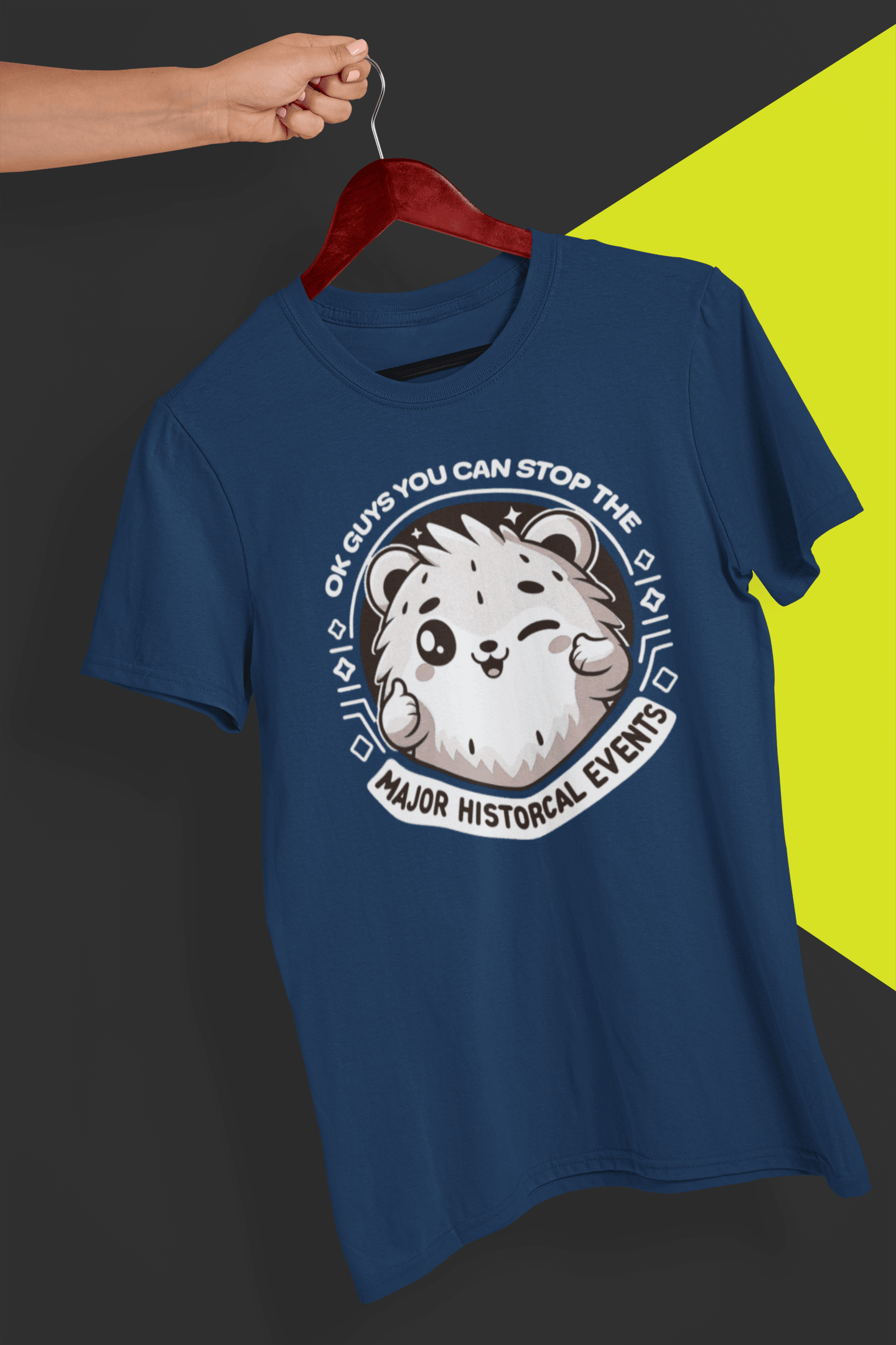 A navy blue T-shirt with a cute hamster doing a thumbs up written &quot;OK GUYS YOU CAN STOP THE MAJOR HISTORICAL EVENTS&quot;. is hung on a red hanger, held by a hand against a split black and yellow background.