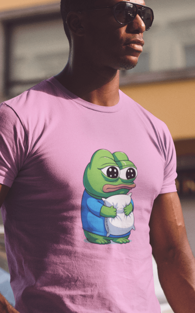 A man wears a pink T-shirt featuring a cute apu apustaja pepe in the middle hugging a pillow.