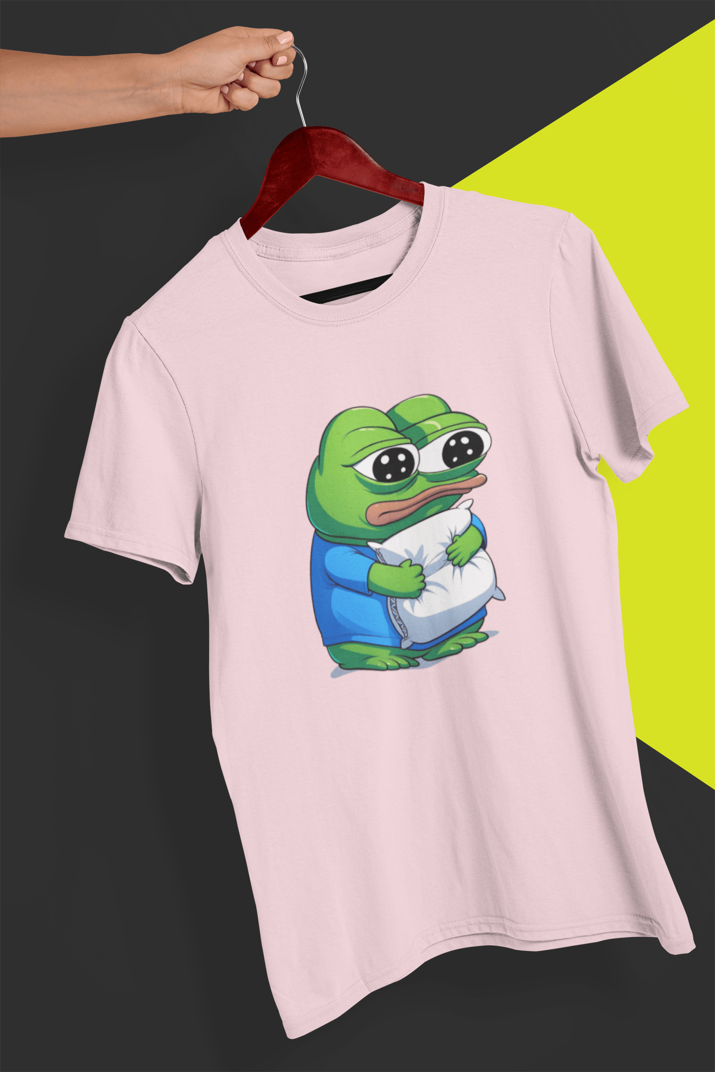 A light pink T-shirt with a cute apu apustaja pepe in the middle hugging a pillow, held by a hand against a split black and yellow background.