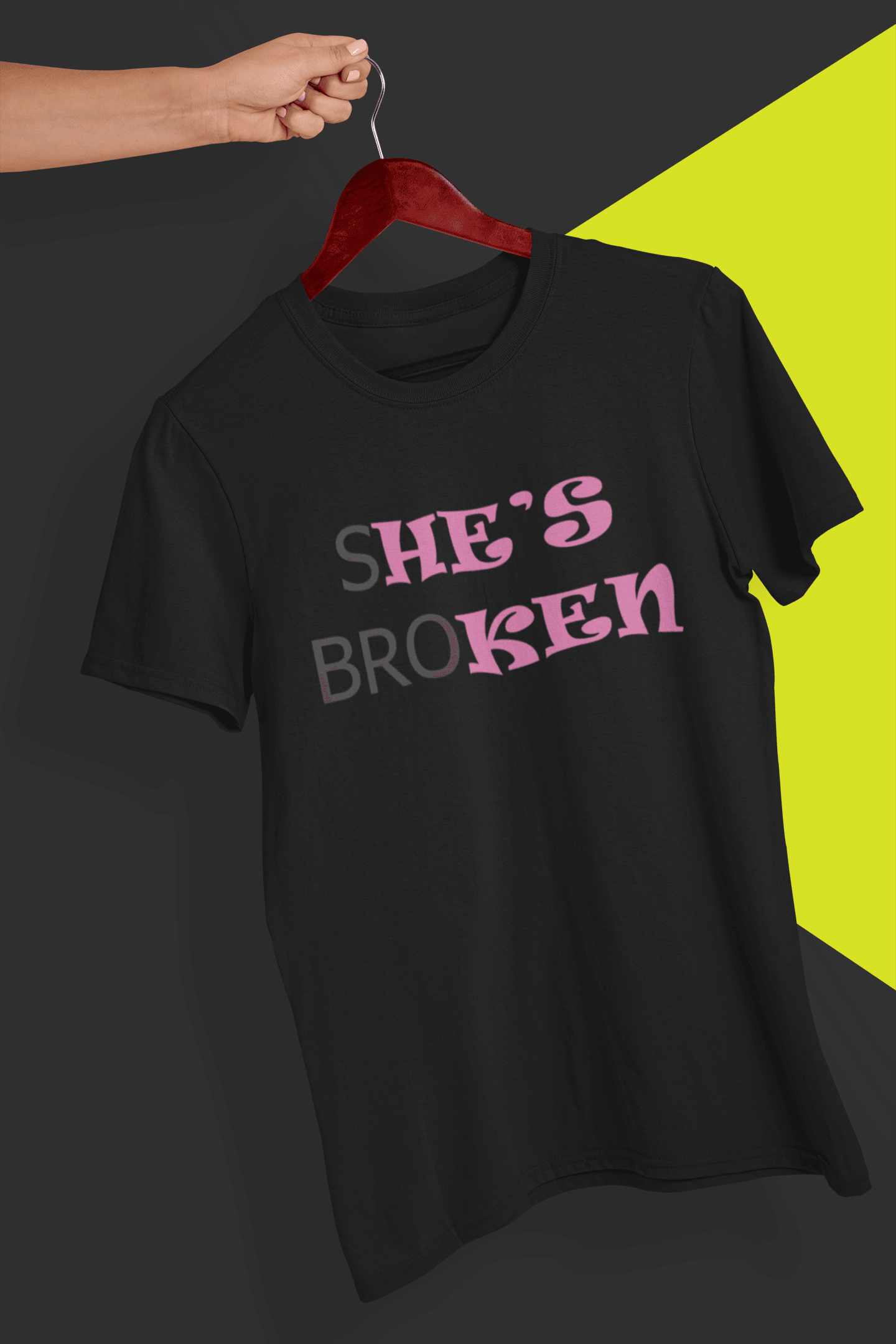 A black T-shirt that reads broth &quot;SHE&#39;S BROKEN&quot; and &quot;HE IS KEN&quot;, in pink and black text; the tshirt is hung on a red hanger, held by a hand against a split black and yellow background.