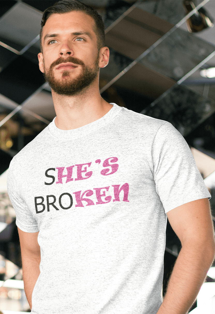 A person in a white T-shirt that reads broth &quot;SHE&#39;S BROKEN&quot; and &quot;HE IS KEN&quot;, in pink and black text, with a contemplative pose.