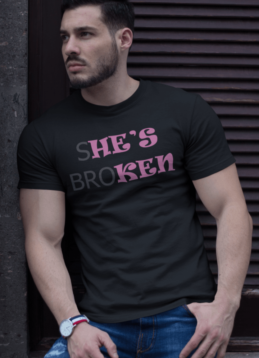 A man in a black T-shirt with the pink and black text that reads broth &quot;SHE&#39;S BROKEN&quot; and &quot;HE IS KEN&quot;, the man is standing in front of a brown corrugated shutter.