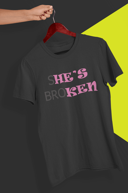 A dark gray T-shirt that reads broth &quot;SHE&#39;S BROKEN&quot; and &quot;HE IS KEN&quot;, in pink and black text; the tshirt is hung on a red hanger, held by a hand against a split black and yellow background.