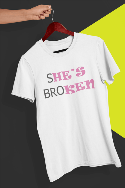 A white T-shirt that reads broth &quot;SHE&#39;S BROKEN&quot; and &quot;HE IS KEN&quot;, in pink and black text; the tshirt is hung on a red hanger, held by a hand against a split black and yellow background.