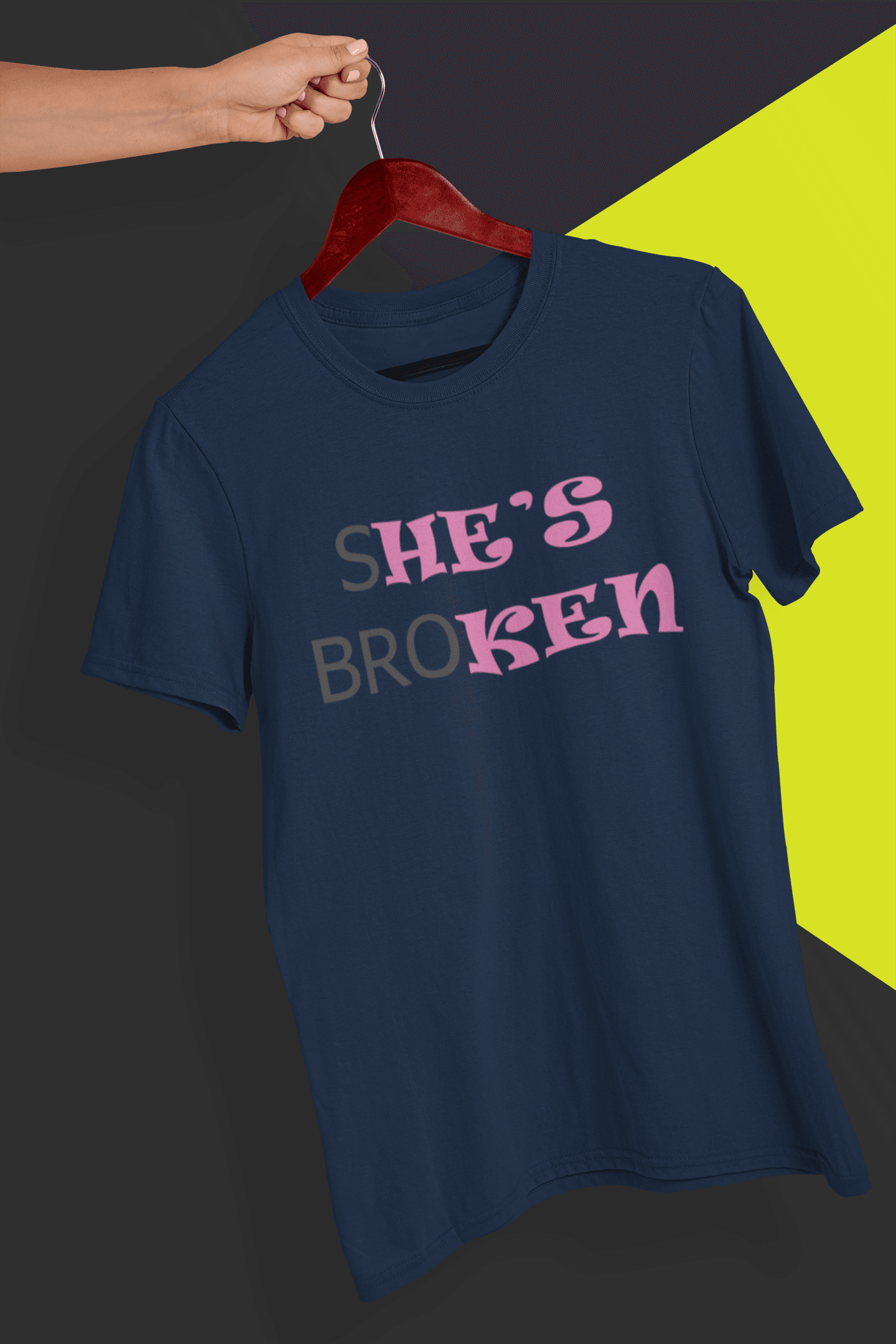 A navy blue T-shirt that reads broth &quot;SHE&#39;S BROKEN&quot; and &quot;HE IS KEN&quot;, in pink and black text; the tshirt is hung on a red hanger, held by a hand against a split black and yellow background.