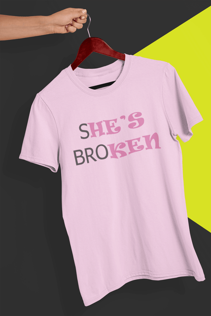 A pink T-shirt that reads broth &quot;SHE&#39;S BROKEN&quot; and &quot;HE IS KEN&quot;, in pink and black text; the tshirt is hung on a red hanger, held by a hand against a split black and yellow background.