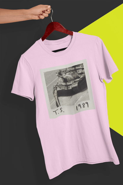 A pink T-shirt with Taylor Swift in front of a tank written &quot;T.S. 1989&quot;, held by a hand against a split black and yellow background.