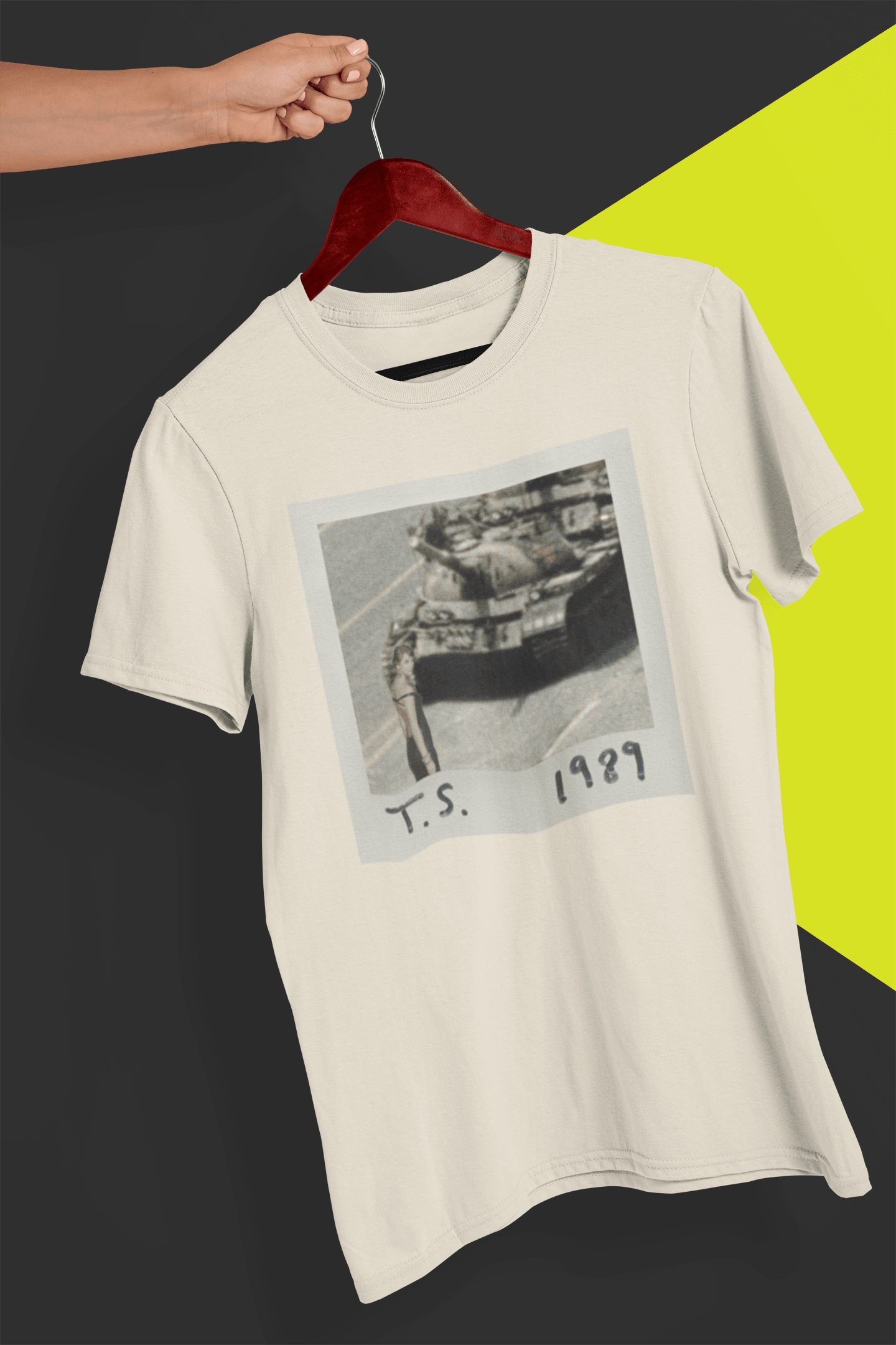 An off-white T-shirt with Taylor Swift in front of a tank written &quot;T.S. 1989&quot;, held by a hand against a split black and yellow background.