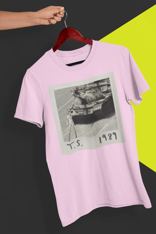 A pink T-shirt with Taylor Swift in front of a tank written &quot;T.S. 1989&quot;, held by a hand against a split black and yellow background.