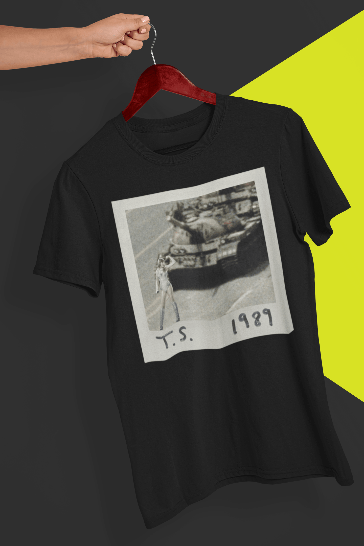 A black T-shirt with Taylor Swift in front of a tank written &quot;T.S. 1989&quot;, held by a hand against a split black and yellow background.