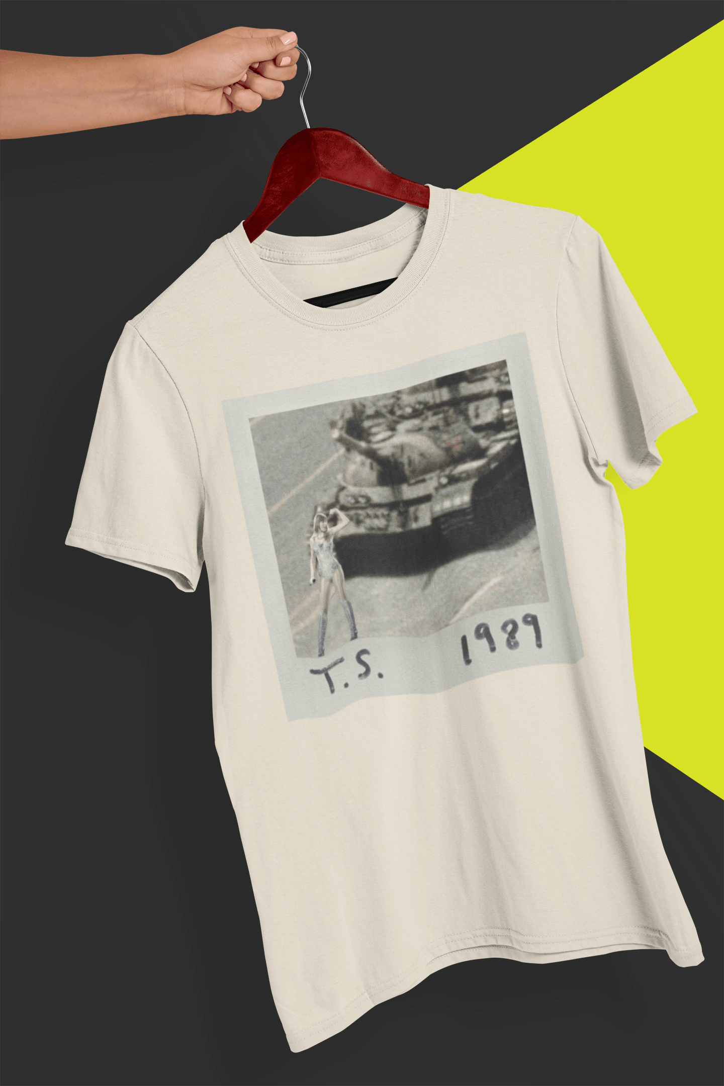 An off-white T-shirt with Taylor Swift in front of a tank written &quot;T.S. 1989&quot;, held by a hand against a split black and yellow background.