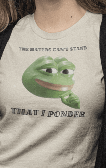 A woman wears a off-white T-shirt featuring a funny 3d smug pepe in the middle, saying &quot;The haters cant&#39;s stand that I ponder&quot;.