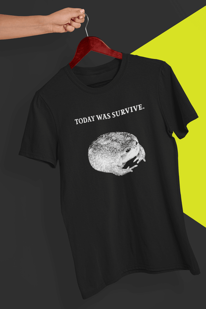 A black T-shirt featuring a cute toad, saying &quot;today was survive.&quot;, is hung on a red hanger, held by a hand against a split black and yellow background.