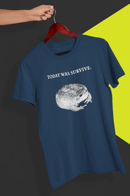 A navy T-shirt featuring a cute toad, saying &quot;today was survive.&quot;, is hung on a red hanger, held by a hand against a split black and yellow background.