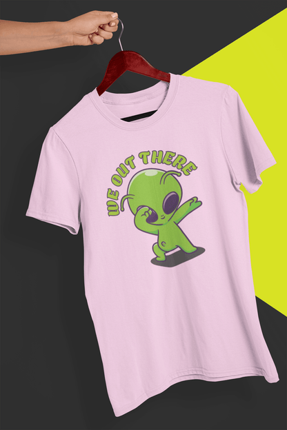 A pink T-shirt with a green alien design and &quot;WE OUT THERE&quot; text is hung on a red hanger, held by a hand against a split black and yellow background.