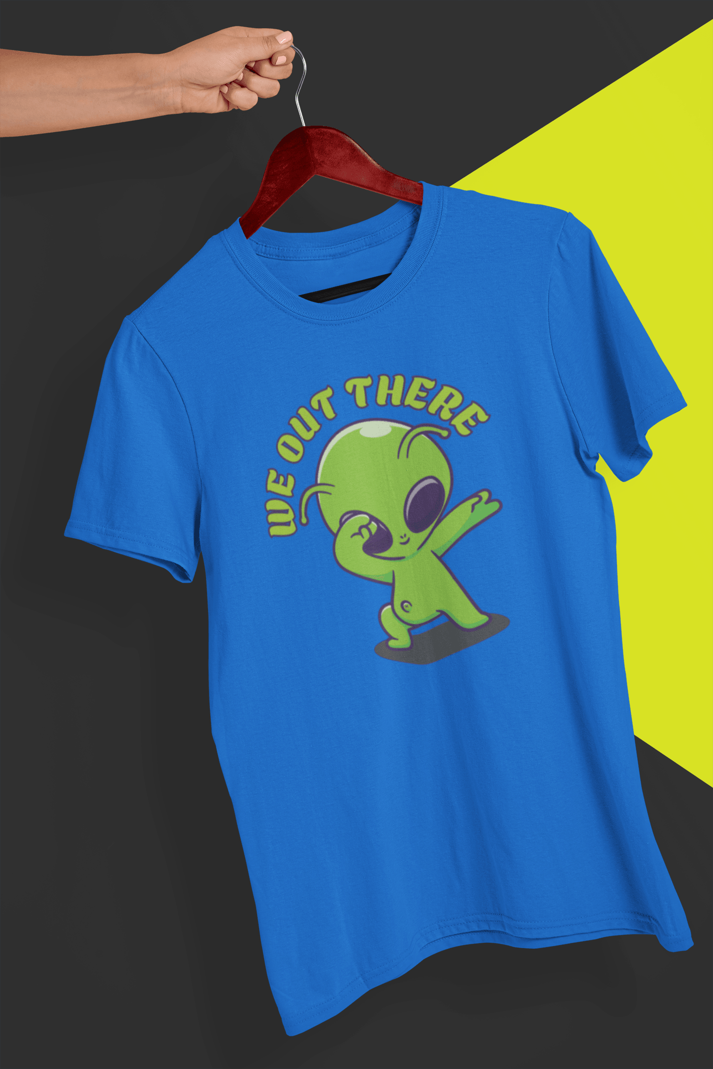 A blue T-shirt with a green alien design and &quot;WE OUT THERE&quot; text is hung on a red hanger, held by a hand against a split black and yellow background.