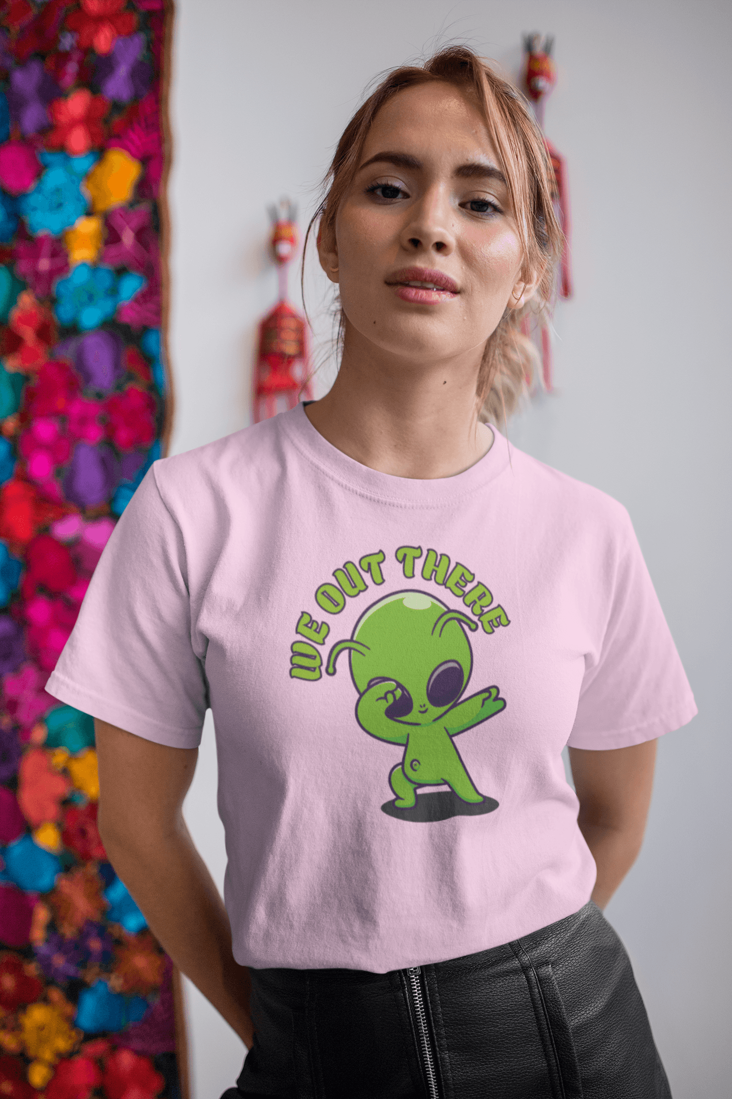 A person wears a pink T-shirt featuring a green cartoon alien with large black eyes making &quot;dab&quot; with the phrase &quot;WE OUT THERE&quot; in bold letters.