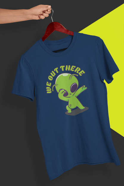 A navy blue T-shirt with a green alien design and &quot;WE OUT THERE&quot; text is hung on a red hanger, held by a hand against a split black and yellow background.