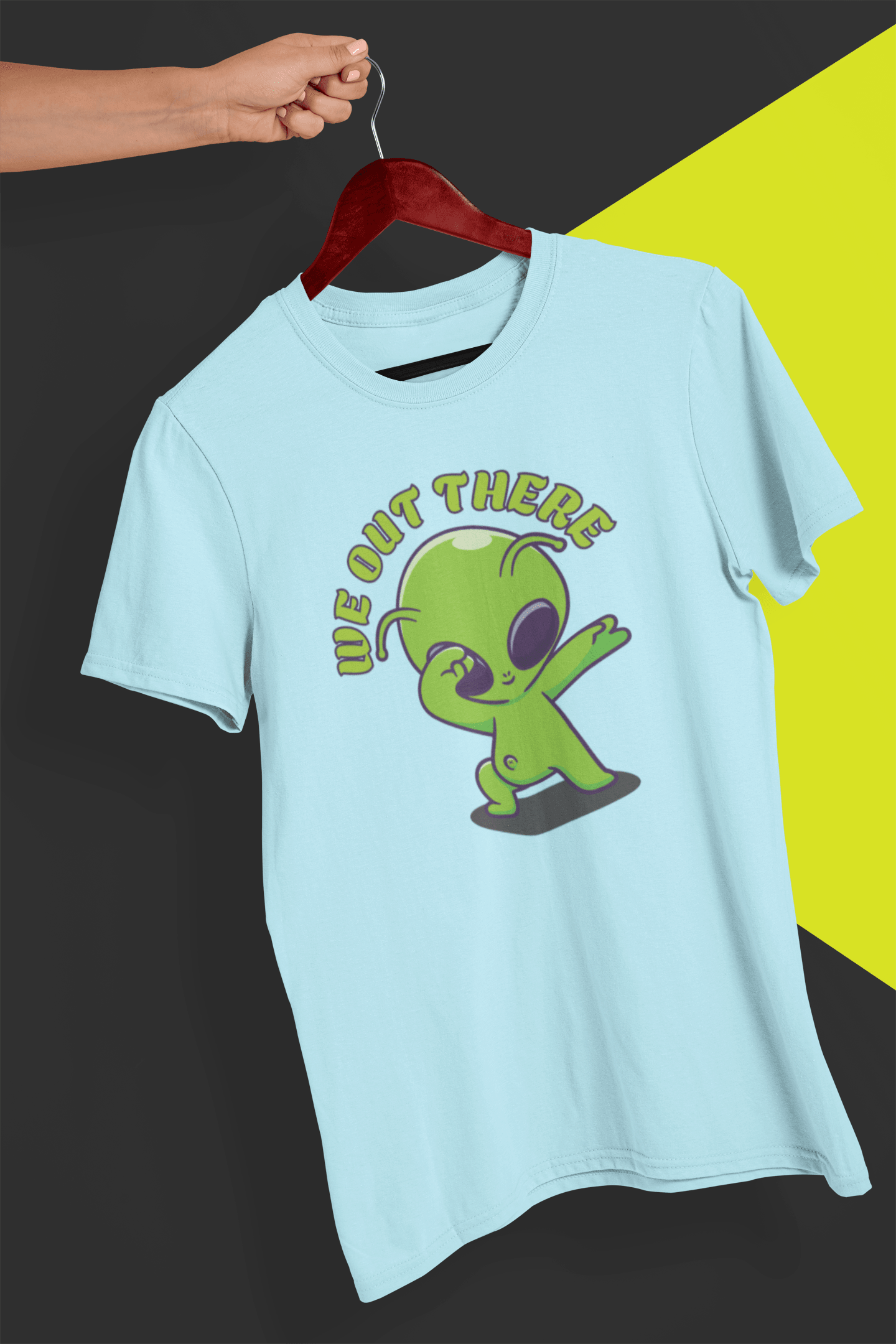 A light blue T-shirt with a green alien design and &quot;WE OUT THERE&quot; text is hung on a red hanger, held by a hand against a split black and yellow background.