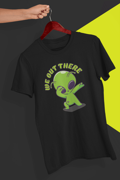 A black T-shirt with a green alien design and &quot;WE OUT THERE&quot; text is hung on a red hanger, held by a hand against a split black and yellow background.