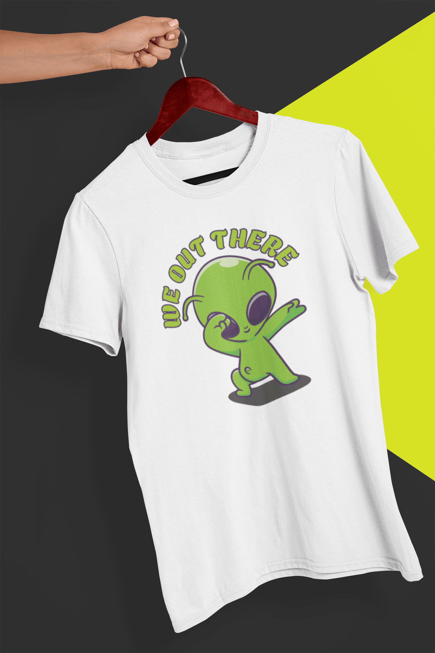 A white T-shirt with a green alien design and &quot;WE OUT THERE&quot; text is hung on a red hanger, held by a hand against a split black and yellow background.