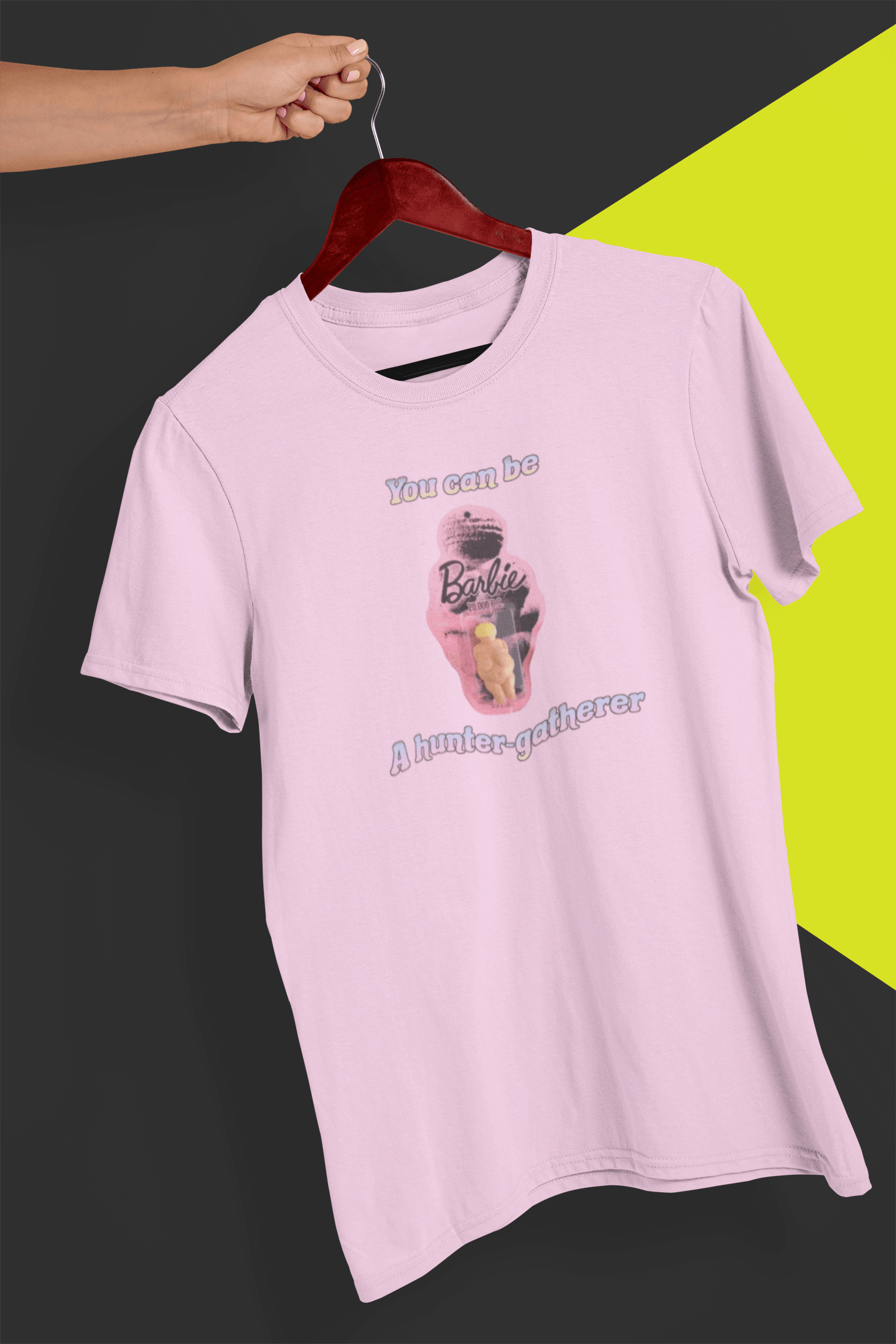 A pink T-shirt featuring the venus impudique that says &quot;Barbie You can be a hunter-gatherer&quot;, held by a hand against a split black and yellow background.